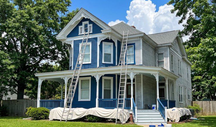 Crew ladders against a blue Victorian house being painted in summer