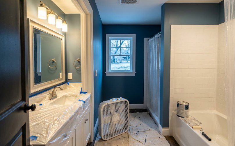Box fan on the floor of a bathroom being painted dark blue, with snow outside the window